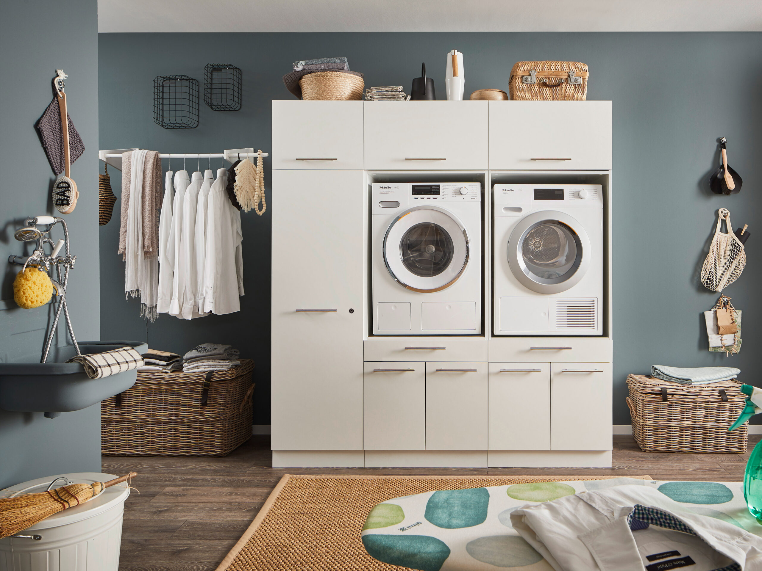 Zweigeräte Wascheinheit Ein Waschraum mit Waschmaschine und Trockner im weißen Möbelmodul, Kleiderstange mit Hemden, Körben und einem Wandwaschtrog.