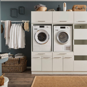 Waschraum mit Zweigeräte Setup Ein Waschraum mit Waschmaschine und Trockner im weißen Möbelmodul, offenen Stauraumfächern, Kleiderstange mit weißen Hemden, Körben und einem Wandwaschtrog.