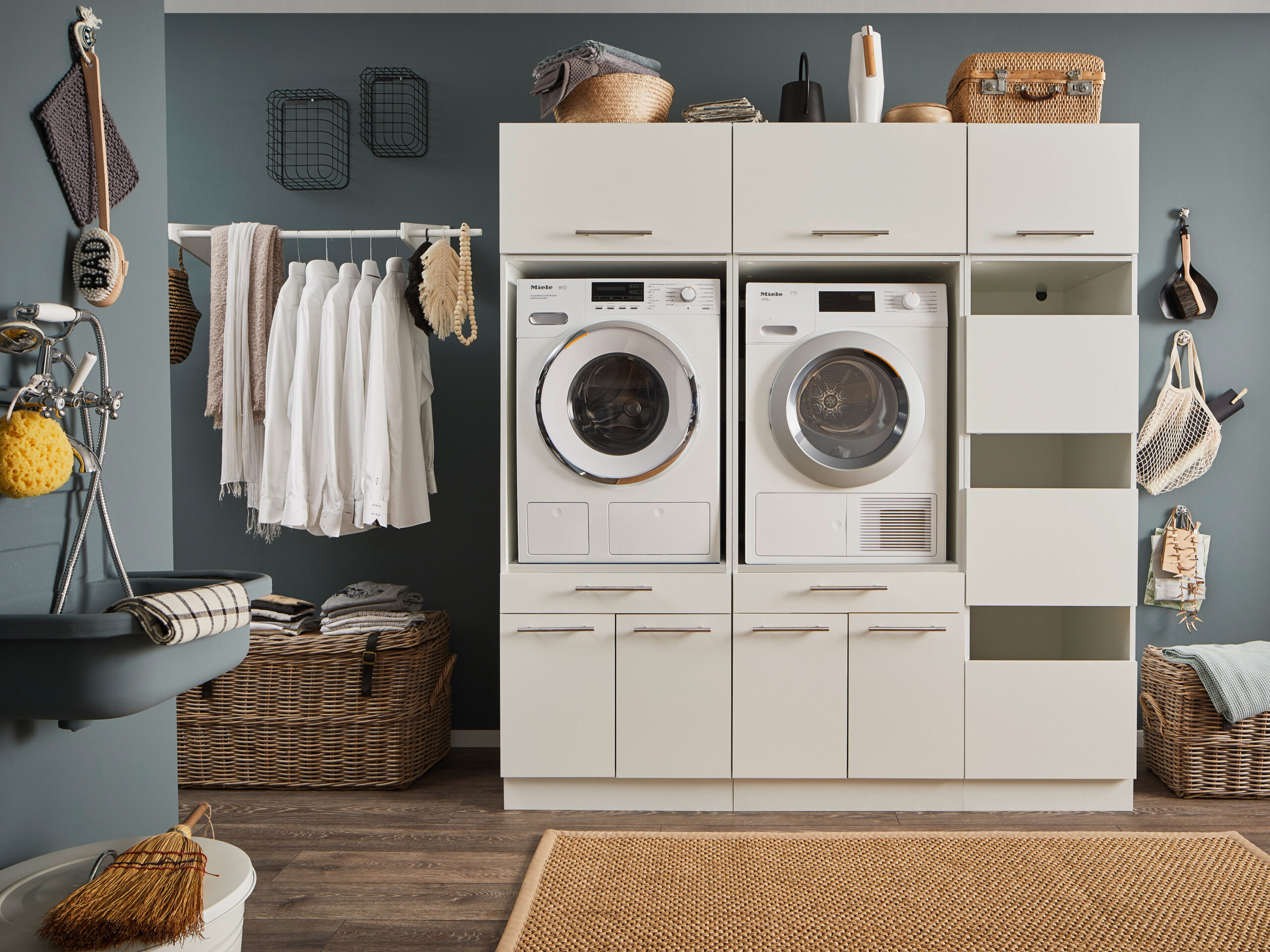 Waschraum mit Zweigeräte Setup Ein Waschraum mit Waschmaschine und Trockner im weißen Möbelmodul, offenen Stauraumfächern, Kleiderstange mit weißen Hemden, Körben und einem Wandwaschtrog.
