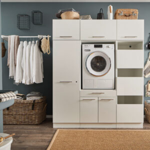 Modulares Waschsystem Ein Waschraum mit eingebauter Waschmaschine im weißen Möbelmodul, offenen Regalfächern, Kleiderstange mit Hemden, Körben und einem Wandwaschtrog.