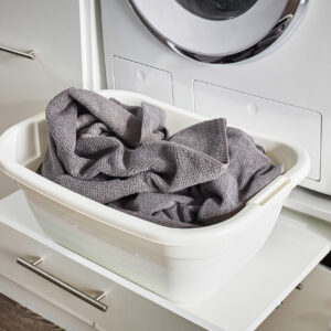 Eine weiße Wanne mit grauen Textilien steht auf einer herausgezogenen Ablage unter einer Waschmaschine in einem hellen Hauswirtschaftsbereich.