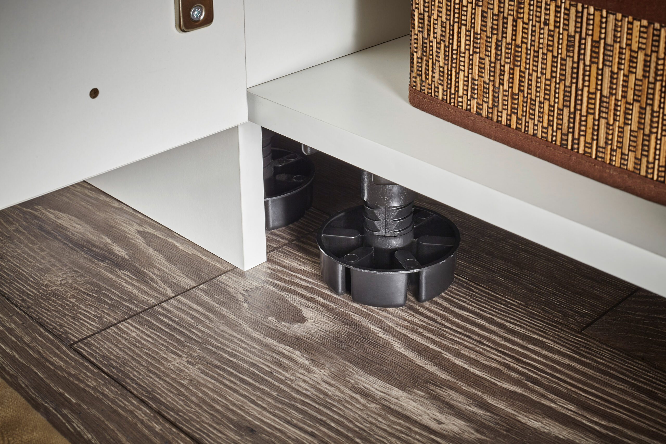 Detailansicht höhenverstellbarer Möbelfüße unter einem weißen Schrank auf Holzboden.