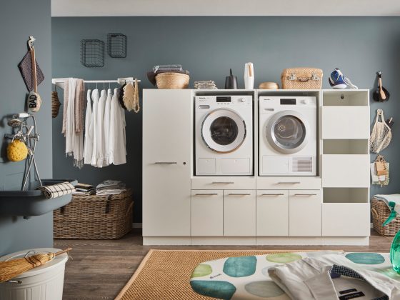 Ein Waschraum mit Waschmaschine und Trockner im wei&szlig;en M&ouml;belmodul, Kleiderstange mit Hemden, offenen Regalf&auml;chern, K&ouml;rben und einem Wandwaschtrog.